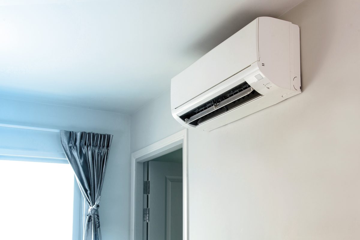 A white wall-mounted heat pump high on a pale blue wall beside a doorway and a window with grey curtains